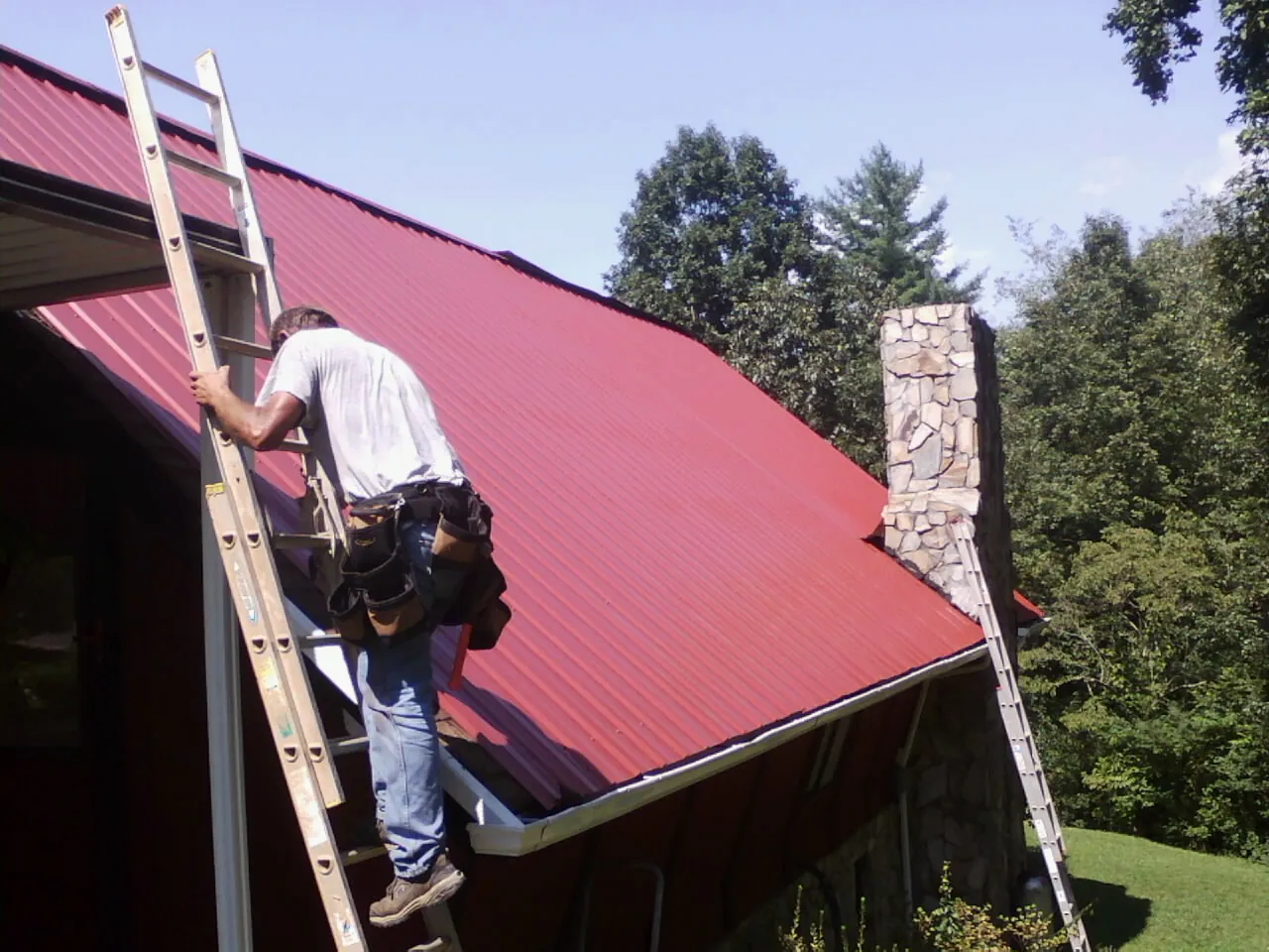 Roofer on a ladder beside a red metal roof and stone chimney — Soffit & Fascia Repair by Roofing Brothers in Townville